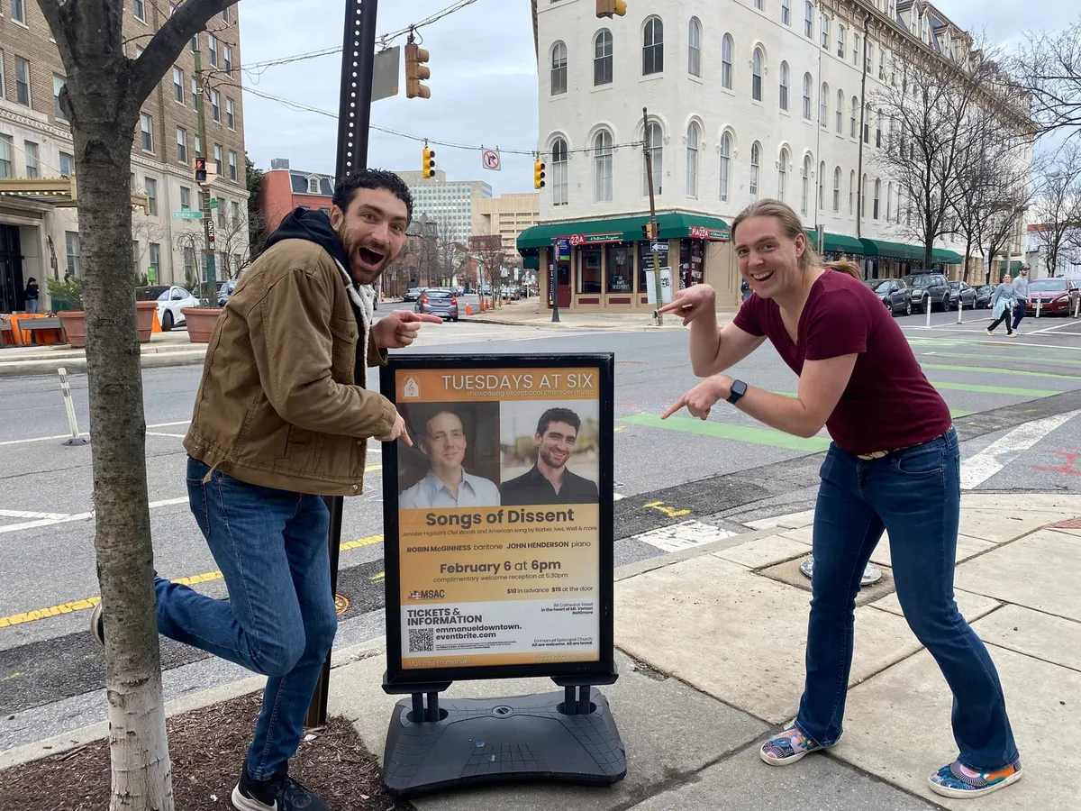 Robin McGinness and John Henderson pointing at a Songs of Dissent concert poster on a Baltimore sidewalk