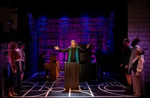 Robin McGinness in The Cradle Will Rock (In Series), arms spread at the podium