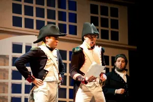 Robin McGinness in bicorne hat and Napoleonic uniform, reading a letter onstage with two fellow performers