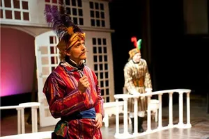 Robin McGinness in a plumed turban and boldly striped robe, in a comic opera scene by a white balustrade